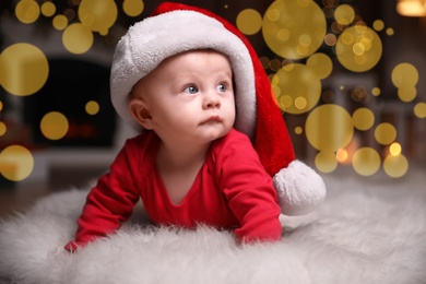 Photo of Cute little baby in red pajamas and Santa hat on floor against blurred festive lights. Christmas suit