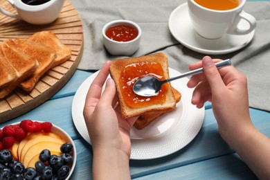 Photo of Woman spreading apricot jam onto tasty toast at light blue wooden table, closeup