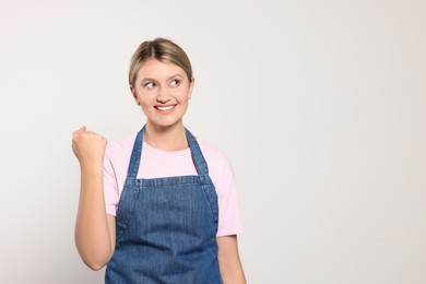 Beautiful young woman in denim apron on light grey background. Space for text