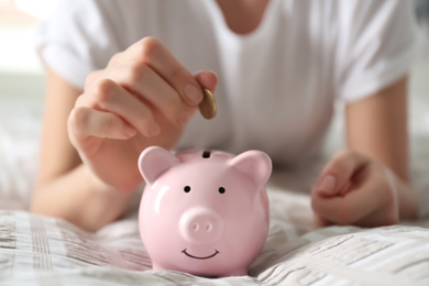 Photo of Woman putting money into piggy bank on bed at home, closeup