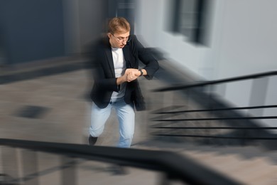 Image of Being late. Young man checking time while running up stairs indoors. Motion blur effect