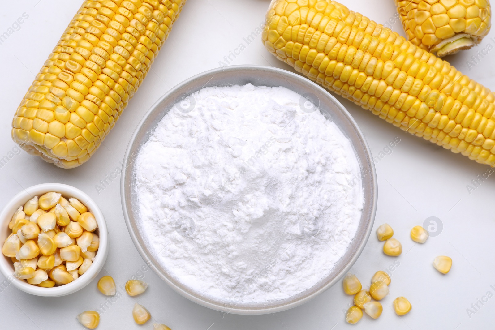 Photo of Bowl with corn starch, ripe cobs and kernels on white table, flat lay