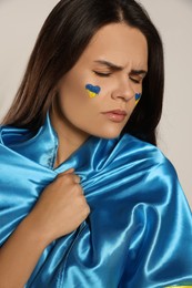 Sad young woman with face paint and Ukrainian flag on beige background