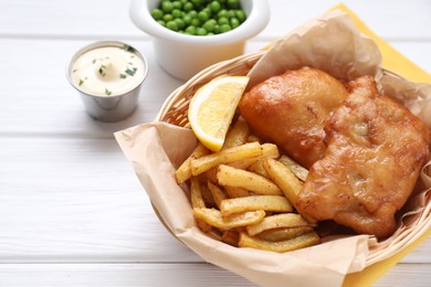 Photo of Tasty fish, chips, sauce and lemon in wicker bowl on white wooden table, closeup. Space for text