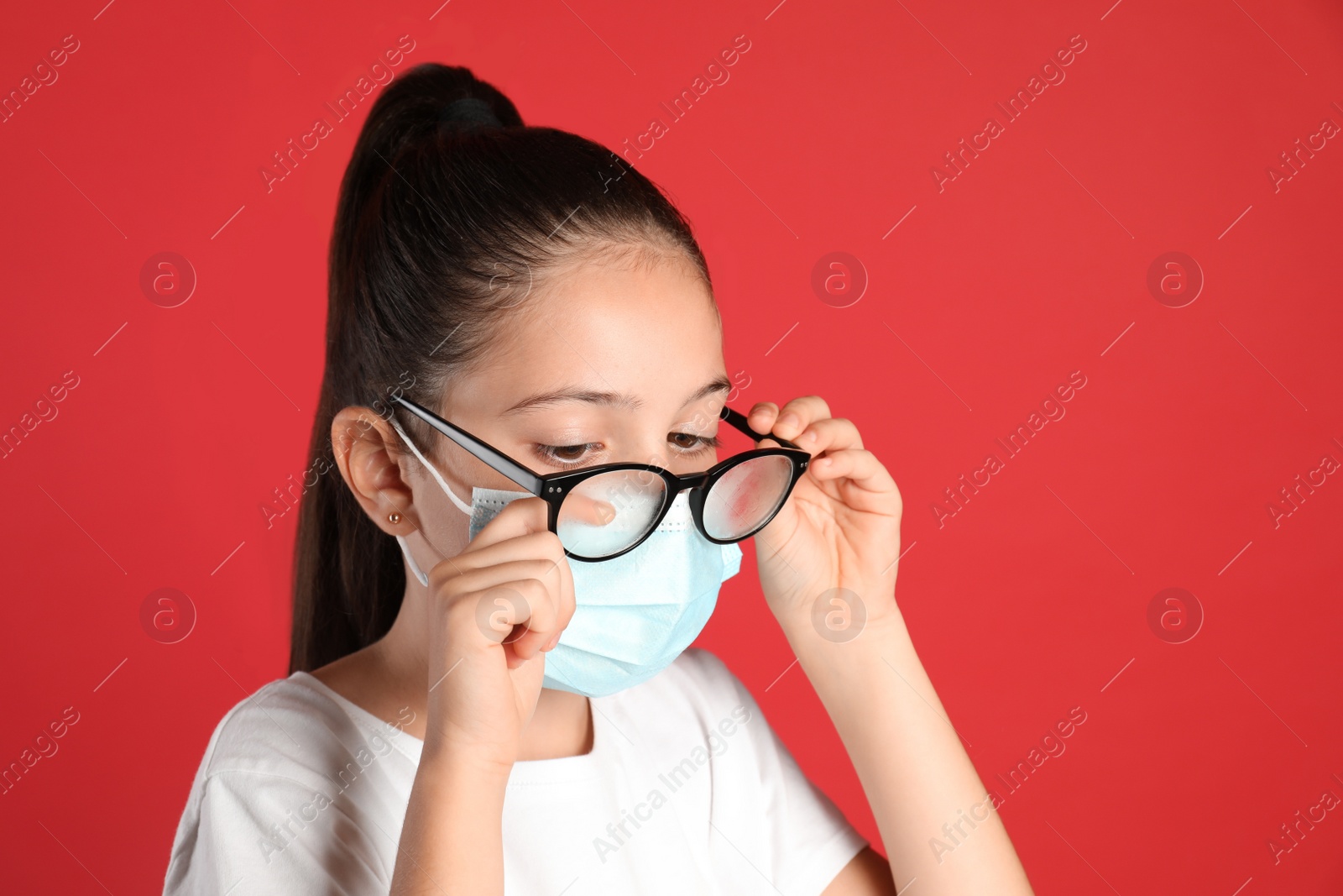 Photo of Little girl wiping foggy glasses caused by wearing medical face mask on red background. Protective measure during coronavirus pandemic