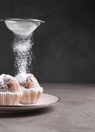 Photo of Woman with sieve sprinkling powdered sugar onto muffins at grey textured table, closeup