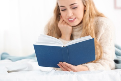 Attractive young woman in cozy warm sweater reading book on bed at home. Space for text