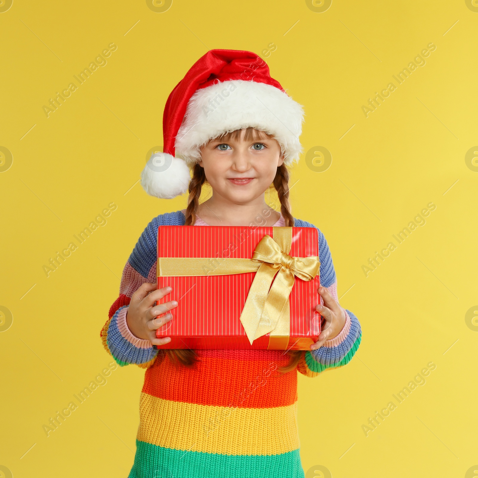 Photo of Cute little girl in Santa hat with Christmas gift on yellow background