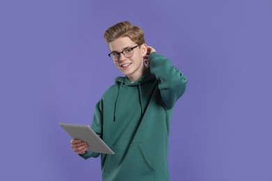 Photo of Teenage boy in stylish eyeglasses using tablet on purple background