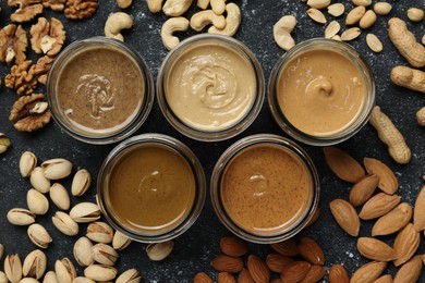Tasty nut butters in jars and raw nuts on dark gray table, flat lay