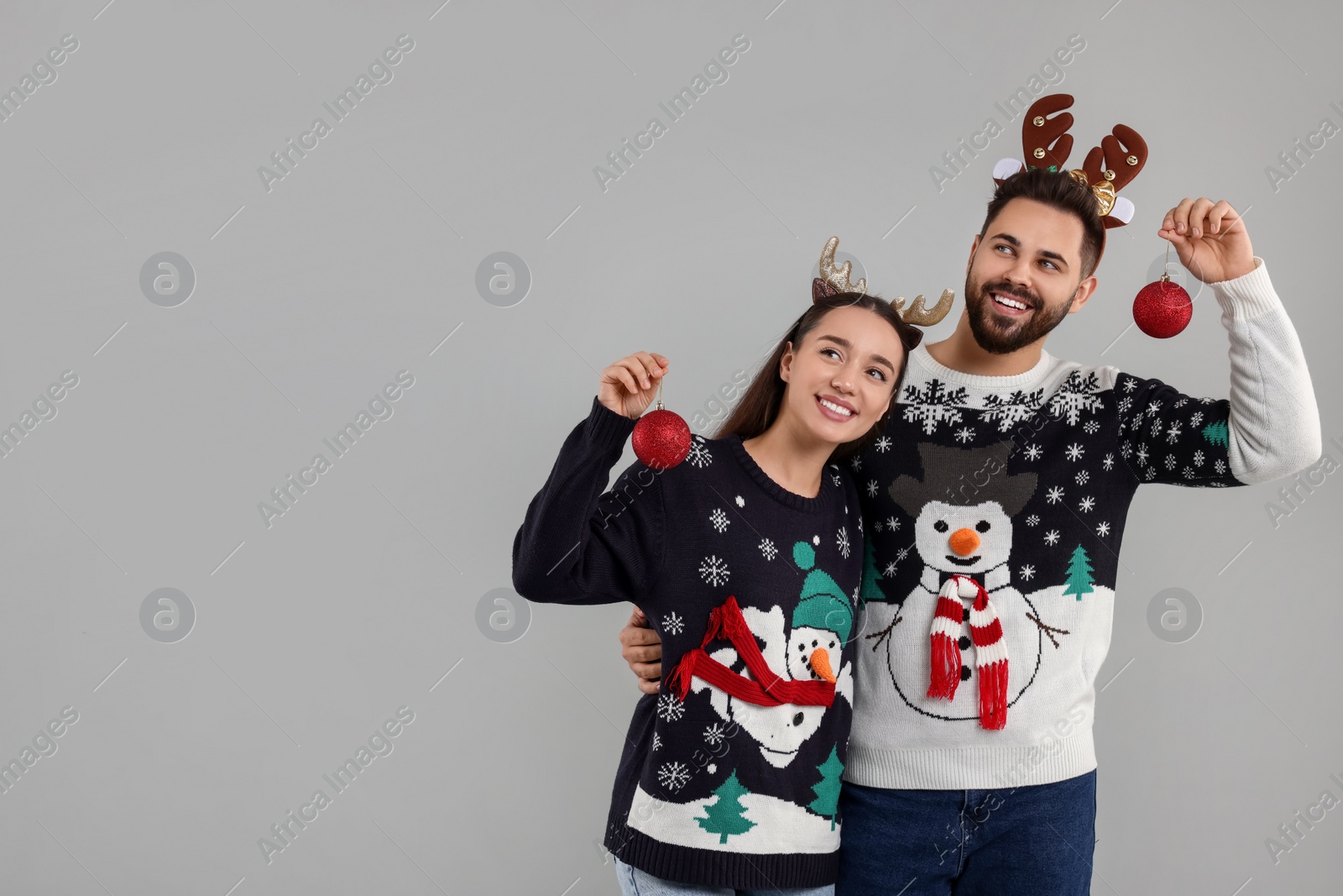 Photo of Happy young couple in Christmas sweaters and reindeer headbands holding festive baubles on grey background. Space for text