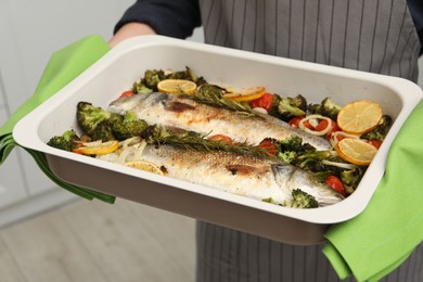 Photo of Woman holding baking dish with delicious fish and vegetables indoors, closeup