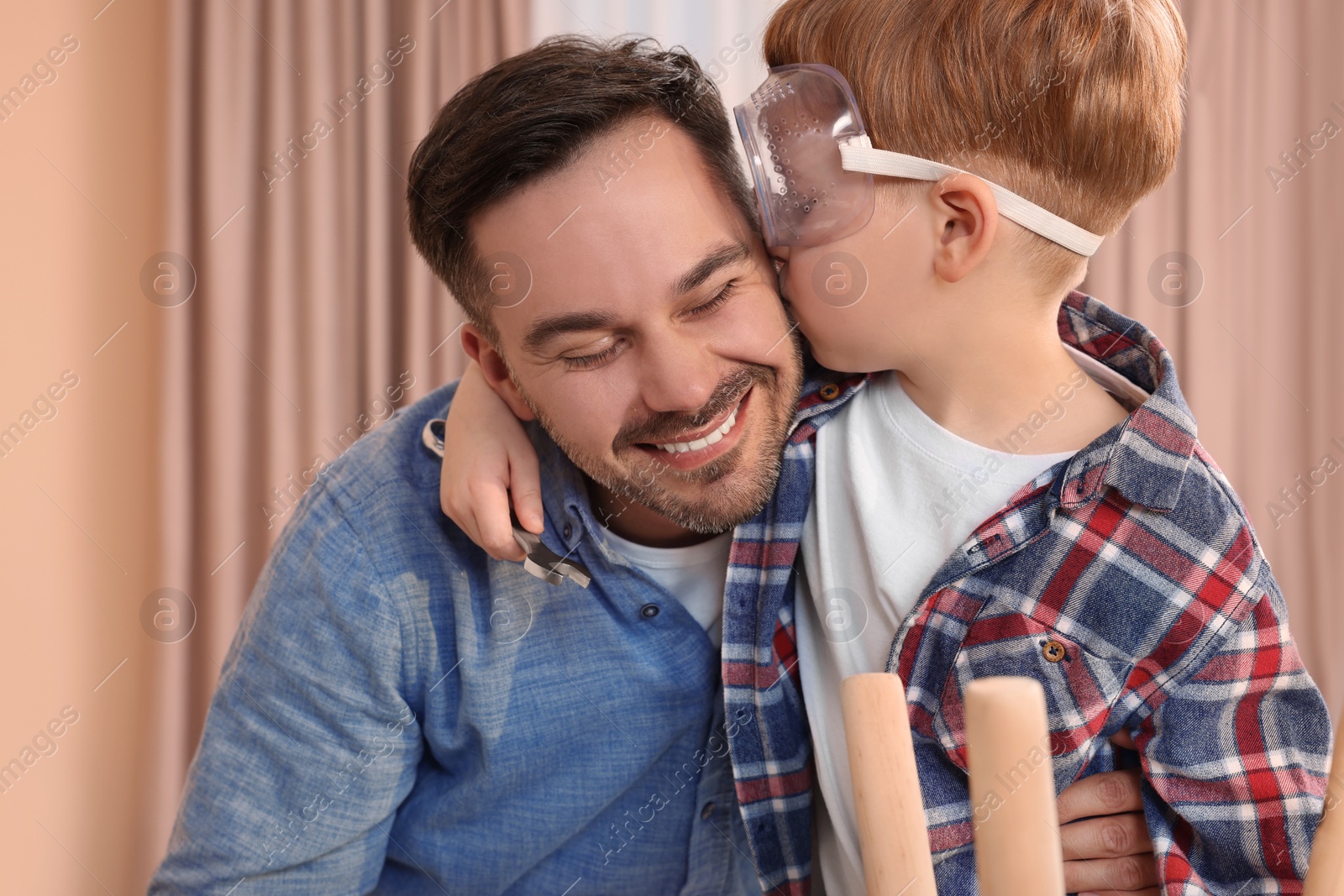 Photo of Son in protective glasses kissing father indoors. Repair work