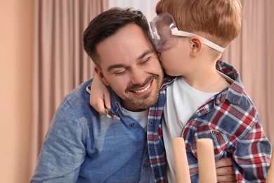 Photo of Son in protective glasses kissing father indoors. Repair work