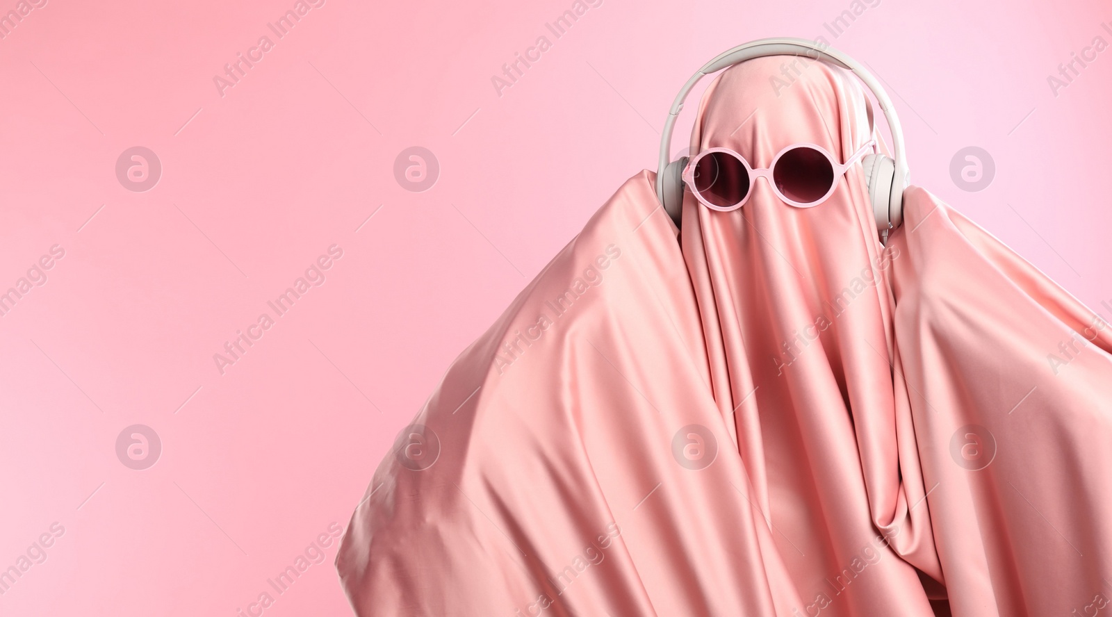 Photo of Glamorous ghost. Woman in sheet with sunglasses and headphones on pink background, space for text