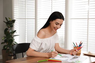Young woman coloring antistress page at table indoors