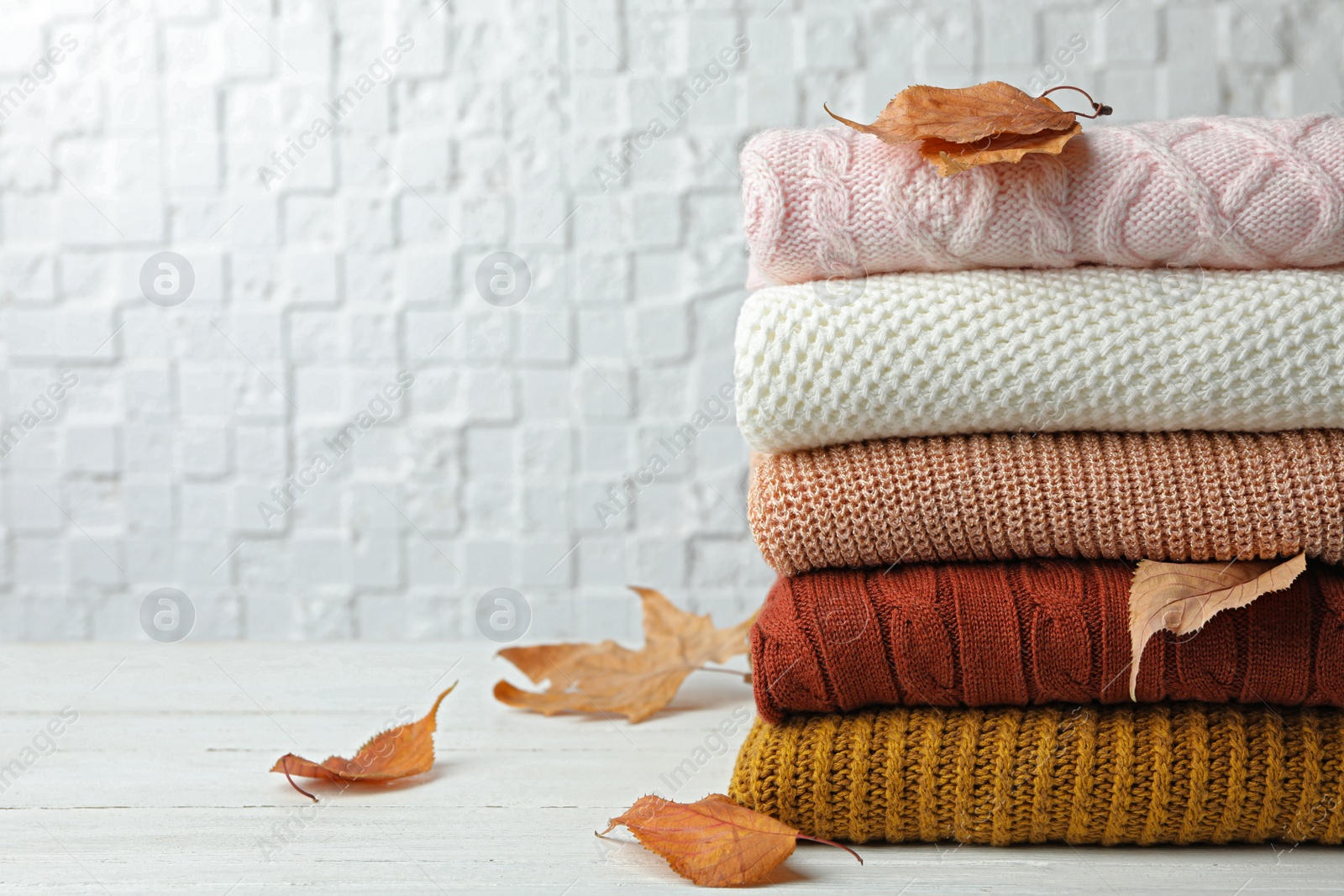 Photo of Stack of warm clothes and autumn leaves on white wooden table against textured wall. Space for text