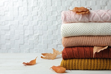 Stack of warm clothes and autumn leaves on white wooden table against textured wall. Space for text