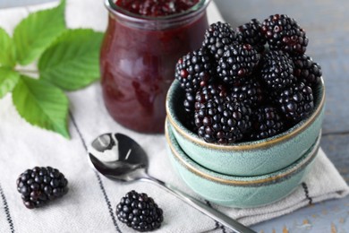 Fresh ripe blackberries, tasty jam and leaves on light grey wooden table, closeup