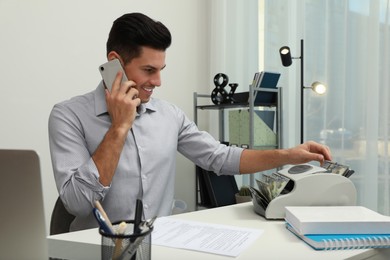 Photo of Man using banknote counter while talking on phone at white table indoors