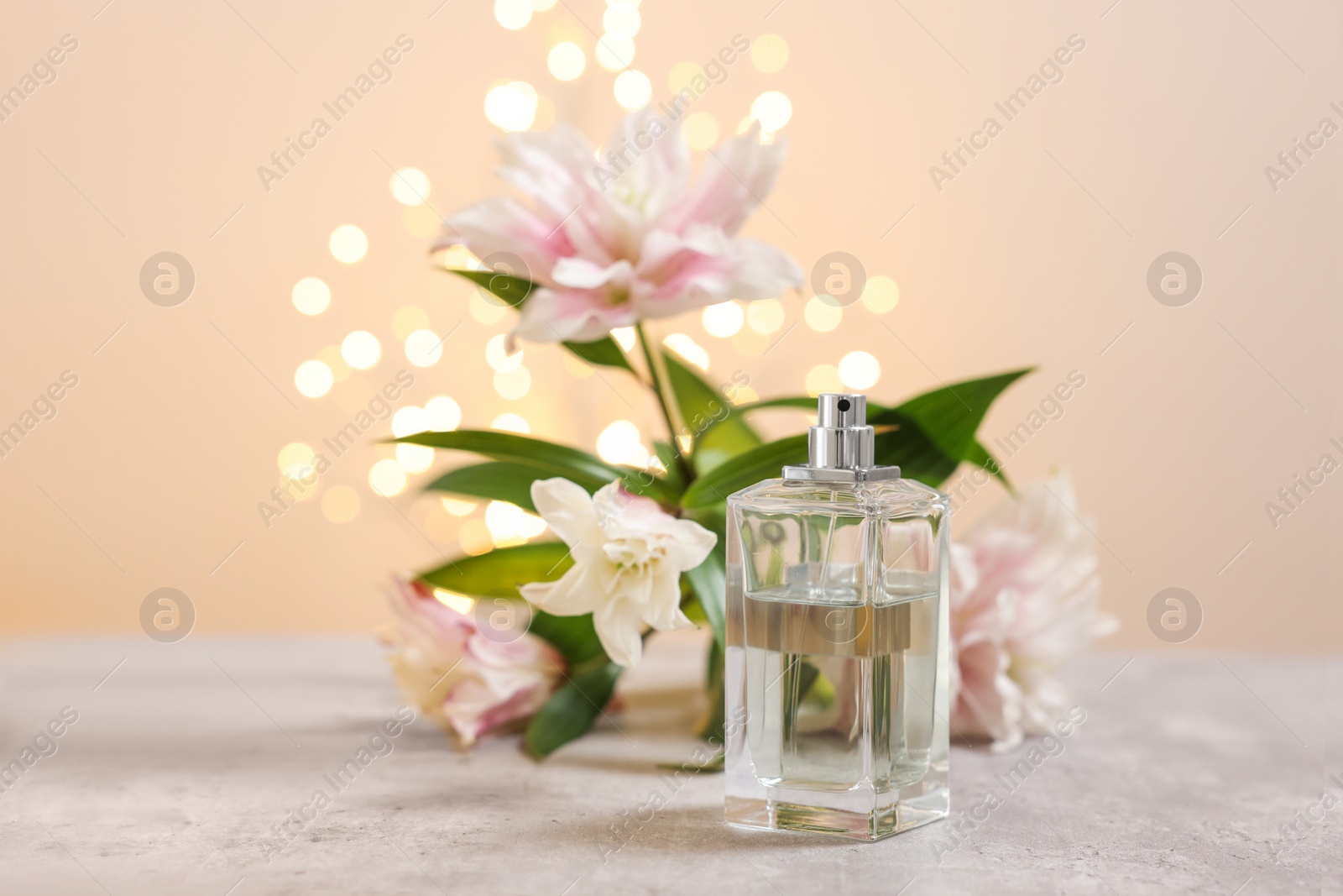 Photo of Bottle of perfume and beautiful lily flowers on table against beige background with blurred lights