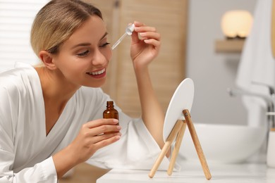 Beautiful woman applying cosmetic serum onto her face in front of mirror at table in bathroom