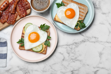 Tasty sandwiches with heart shaped fried eggs for romantic breakfast on white marble table, flat lay