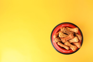 Bone shaped dog cookies in feeding bowl on yellow background, top view. Space for text