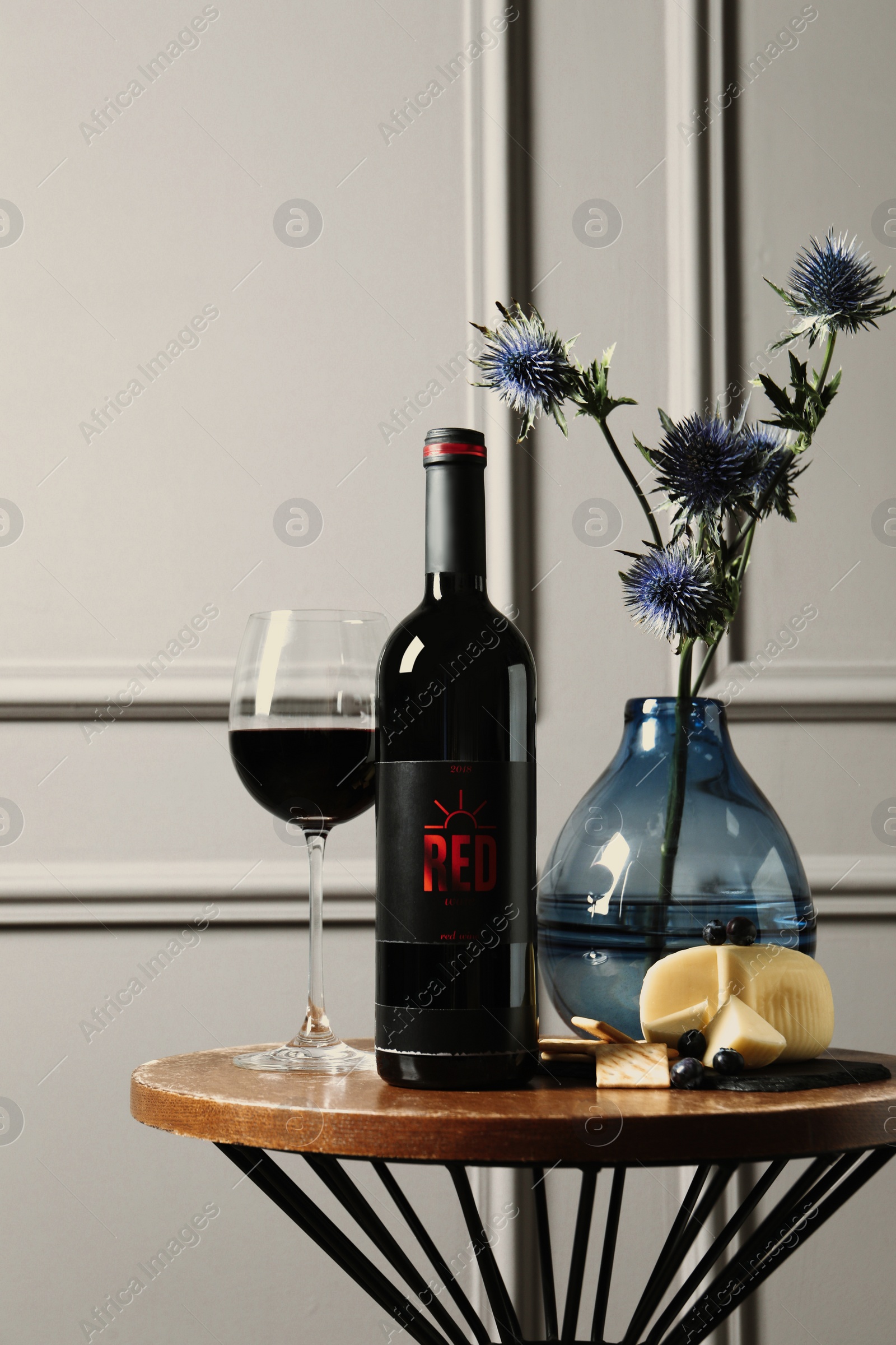 Photo of Bottle of red wine, glass, delicious snacks and beautiful bouquet on wooden table indoors