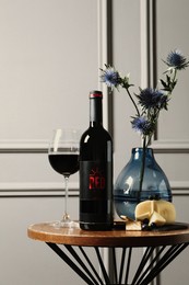 Photo of Bottle of red wine, glass, delicious snacks and beautiful bouquet on wooden table indoors