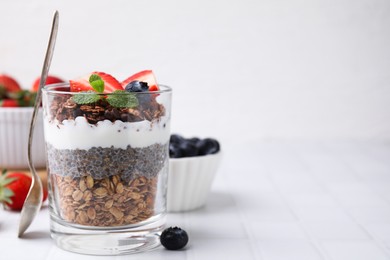 Photo of Tasty granola with berries, yogurt and chia seeds in glass on white tiled table, space for text