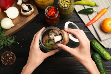Photo of Woman putting champignon into glass jar at black wooden table, top view. Pickling mushrooms