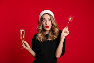 Photo of Surprised young woman wearing Santa hat with glass of champagne and sparkler on red background. Christmas celebration