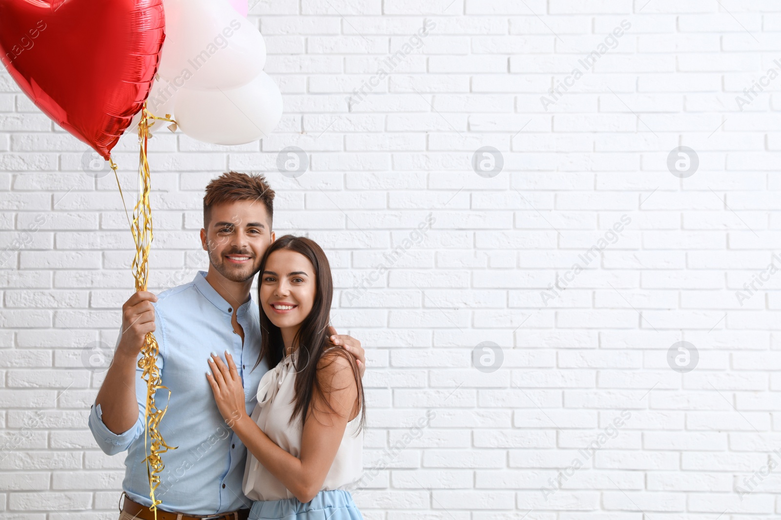 Photo of Young couple with air balloons near white brick wall. Celebration of Saint Valentine's Day