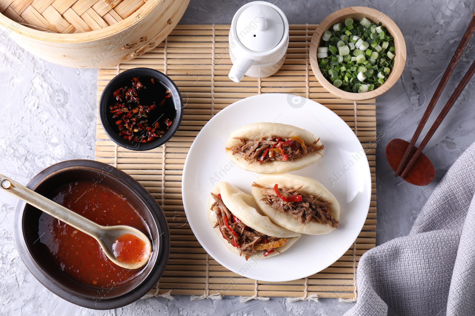 Photo of Delicious gua bao (pork belly buns), chopsticks, green onion and sauces on grey textured table, flat lay