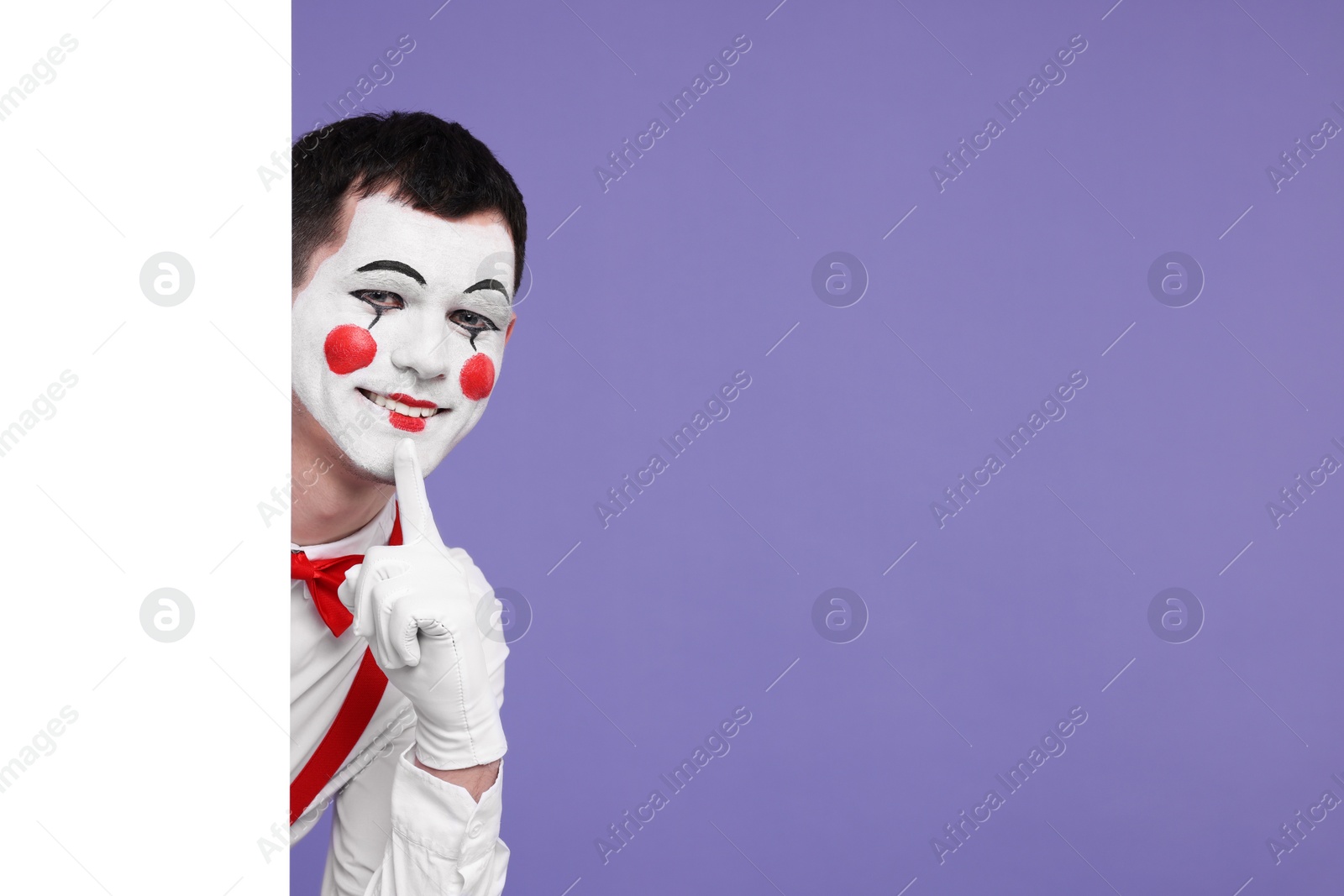 Photo of Funny mime artist peeking out of blank poster on purple background. Space for text