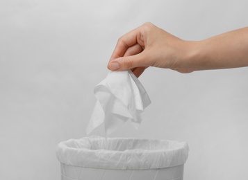 Woman putting paper tissue into trash bin on light background, closeup