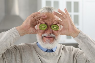 Senior man covering eyes with halves of kiwi indoors