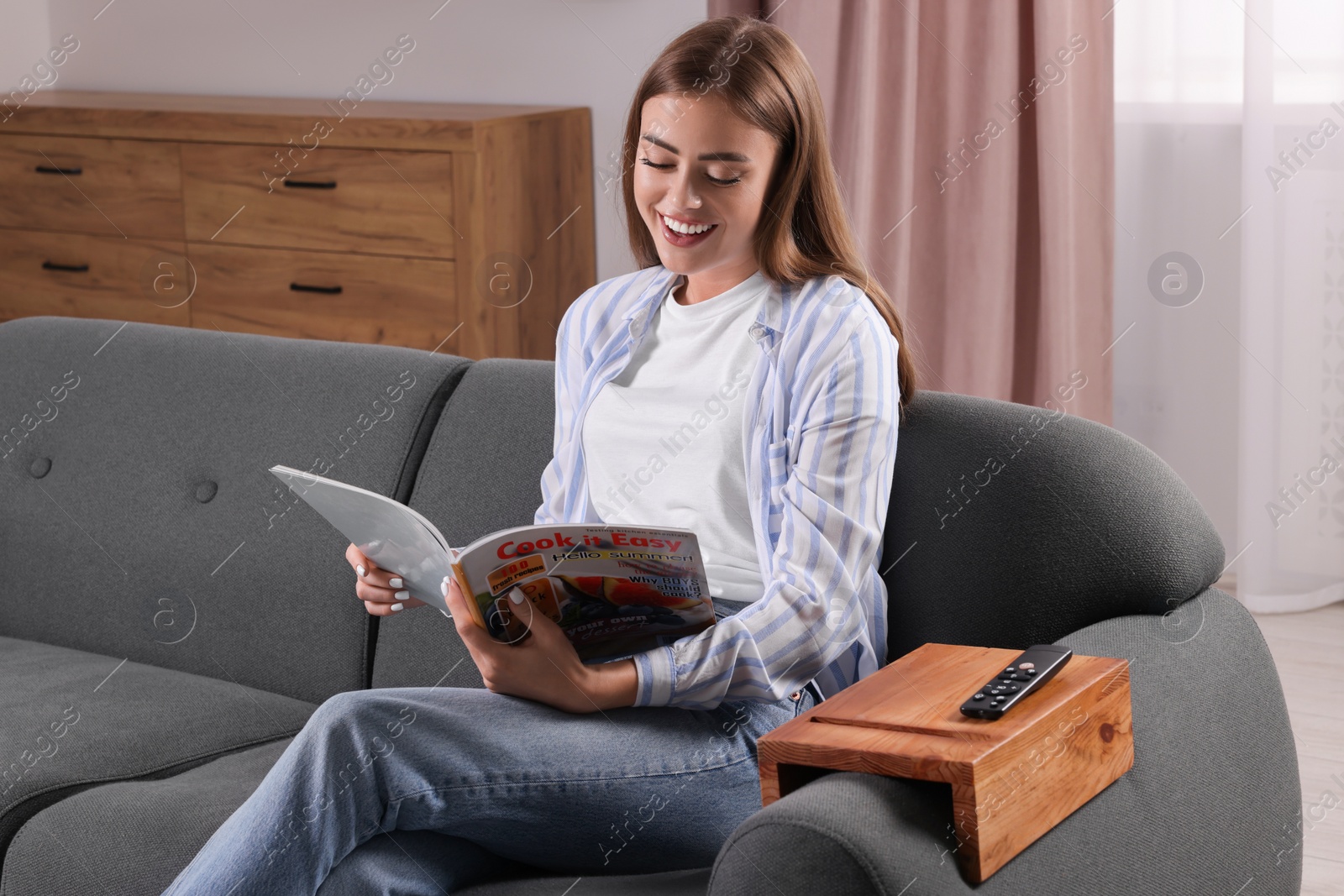 Photo of Happy woman reading magazine on sofa with wooden armrest table at home