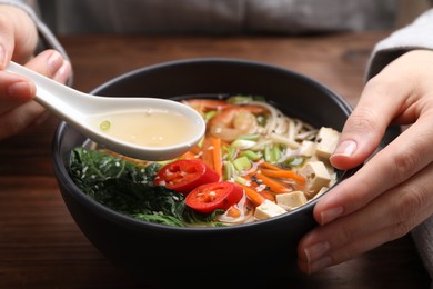 Woman eating delicious ramen with spoon at wooden table, closeup. Noodle soup