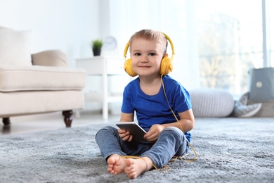 Photo of Cute child with headphones and mobile phone on floor indoors