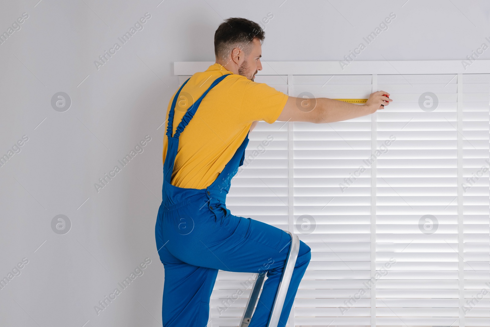 Photo of Worker in uniform using measuring tape while installing horizontal window blinds on stepladder indoors
