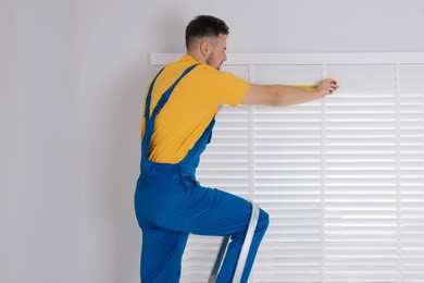 Worker in uniform using measuring tape while installing horizontal window blinds on stepladder indoors