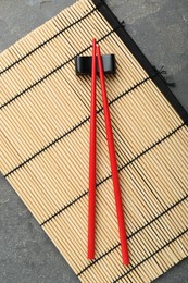 Photo of Bamboo mat with pair of red chopsticks and rest on grey table, top view
