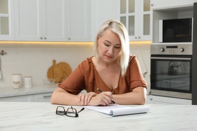 Photo of Middle aged woman solving sudoku puzzle at table in kitchen