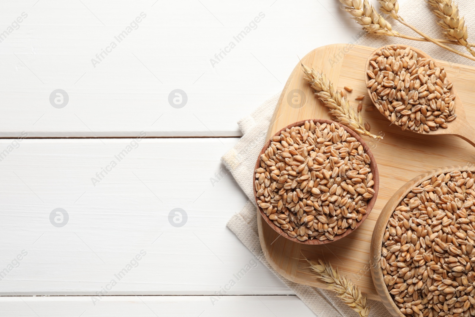 Photo of Wheat grains in bowls and spoon on white wooden table, flat lay. Space for text