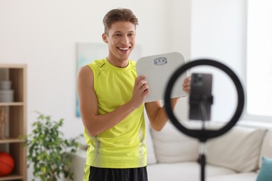Smiling sports blogger holding floor scales while streaming online fitness lesson with phone at home