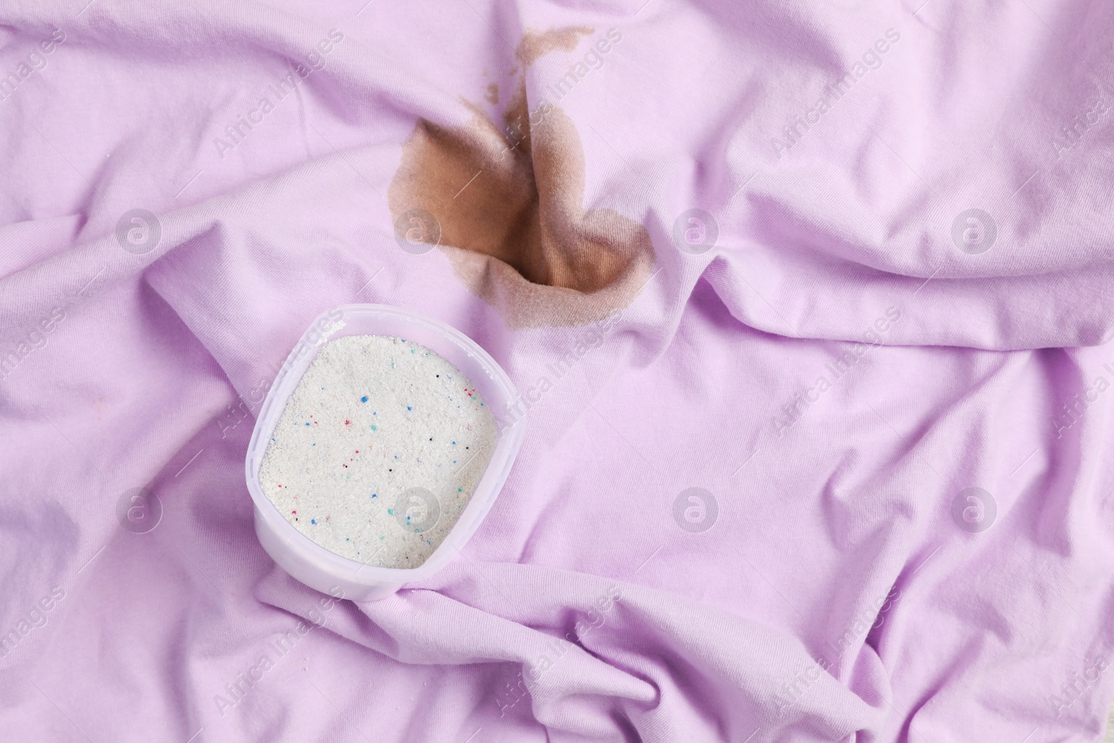 Photo of Measuring cup of powdered detergent on garment with stain, above view. Hand washing laundry