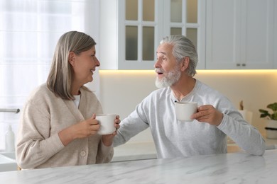 Photo of Affectionate senior couple with cups of drink at white marble table in kitchen