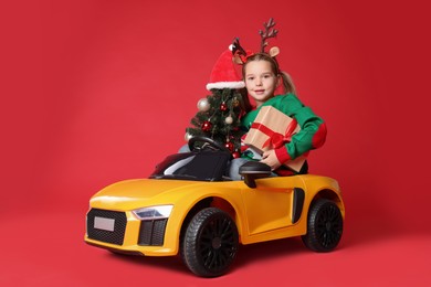 Photo of Cute little girl with Christmas tree and gift box driving children's electric toy car on red background
